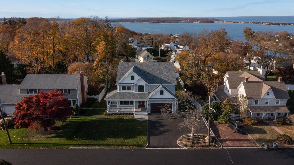 Aerial drone view of a modern two-story gray craftsman home with a large paved driveway in Northport, New York, featuring autumn trees and a scenic background view of the Long Island Sound. The image was taken by Osman Photographics, licensed UAV drone pilot.