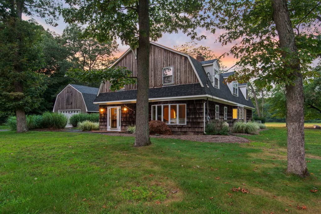 Two-story wood-shingle Dutch colonial house with glowing interior lights, framed by tall oak trees against a pink and orange sunset sky.